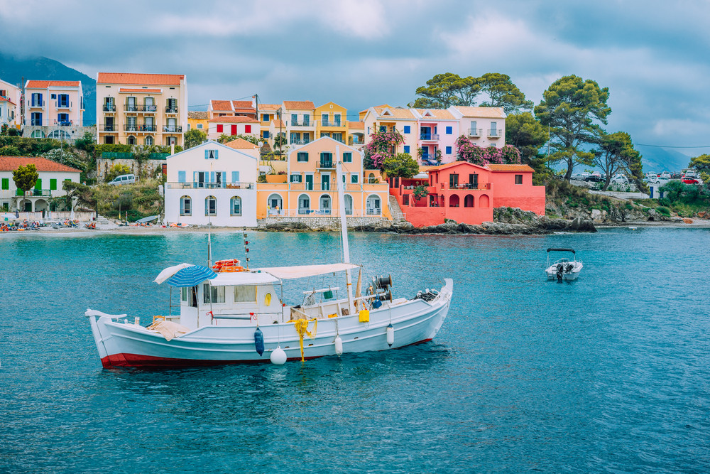 Fishing boat at anchor in blue sea bay of Assos village. Vivid colored houses with clouds in background, Kefalonia island, Greece