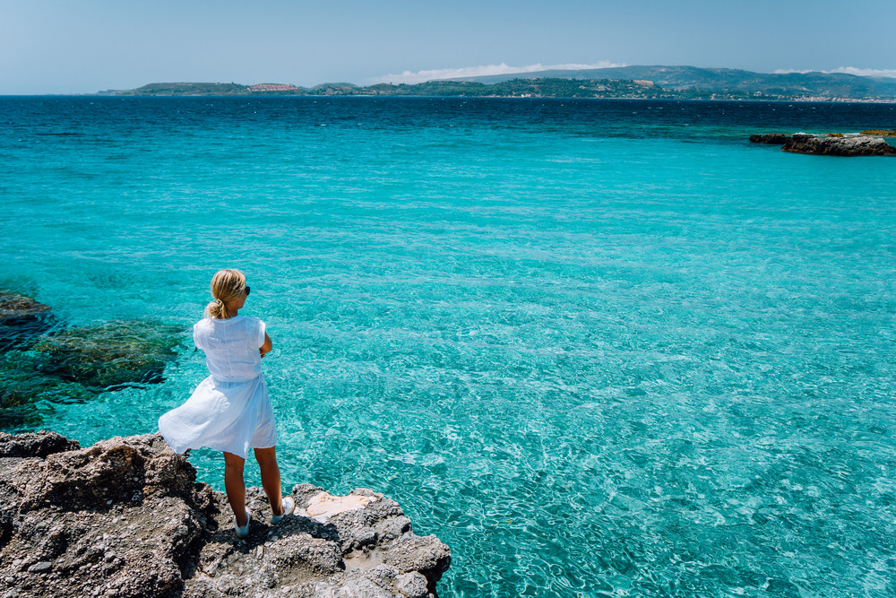 Jung adult women in white dress on summer vacation in front of sea coast landscape of small beach with crystal clear blue azure water (Greece, Kefalonia)