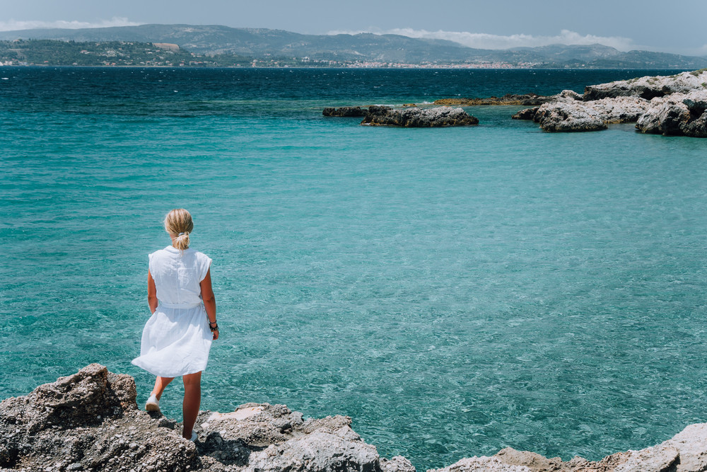 Jung adult women in white dress on summer vacation in front of sea coast landscape of small beach with crystal clear blue water. Greece, Kefalonia