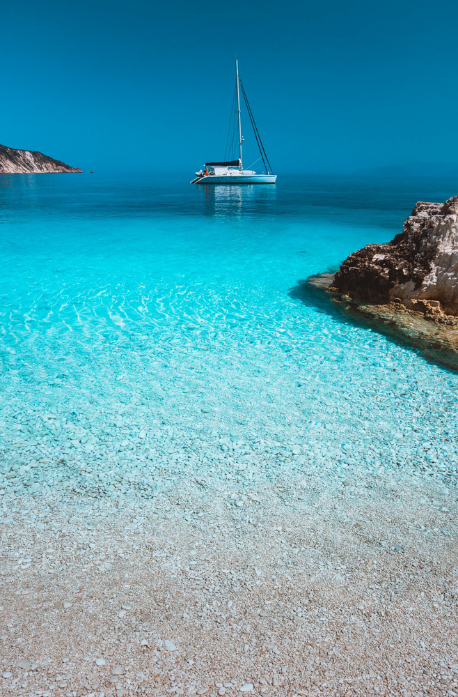 Lonely white sailing catamaran boat drift on calm sea surface. Pure azure clean blue lagoon with shallow water and pebble beach. Some brown rock stones in foreground