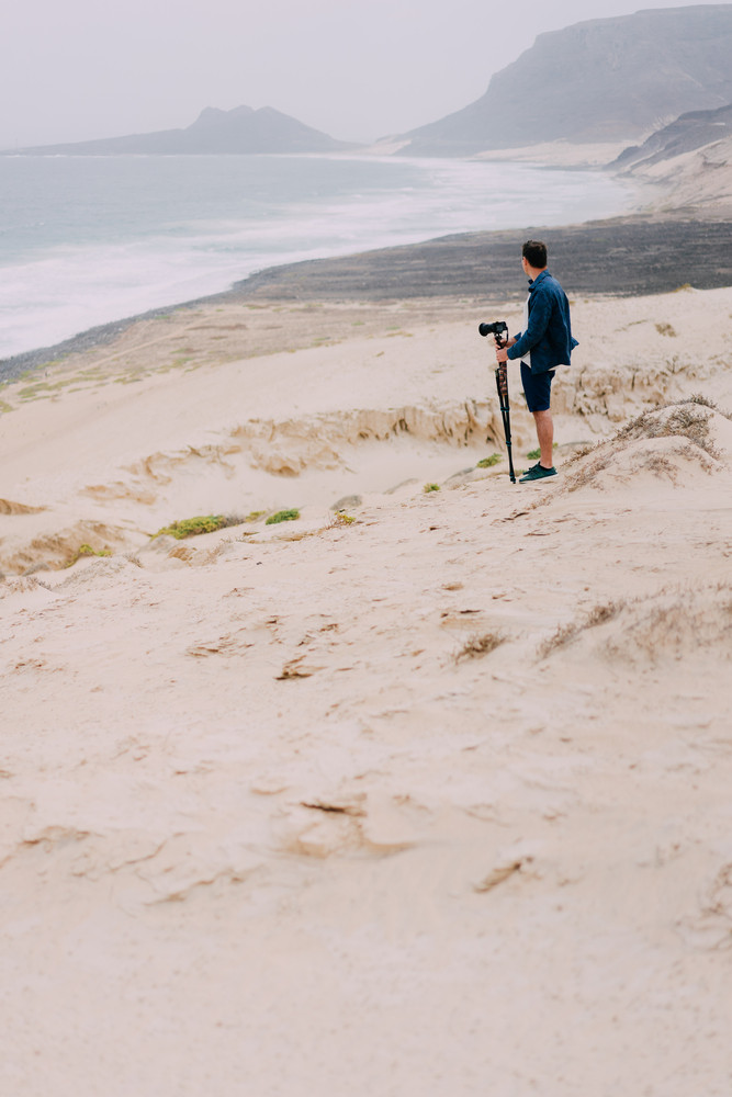 Photographer with camera in desert admitting unique landscape of sand dunes volcanic cliffs on the Atlantic coast. Baia Das Gatas, near Calhau, Sao Vicente Island Cape Verde
