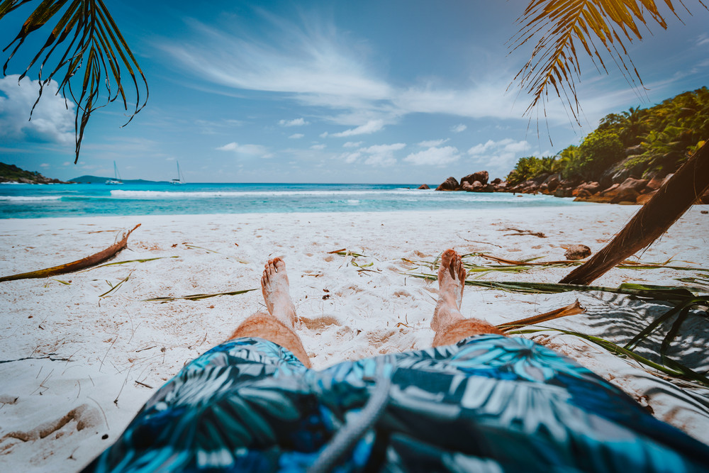 POV shot of man wear blue swimming shorts laying on a beautiful sandy tropical beach Anse Cosos, La Digue, Seychelles