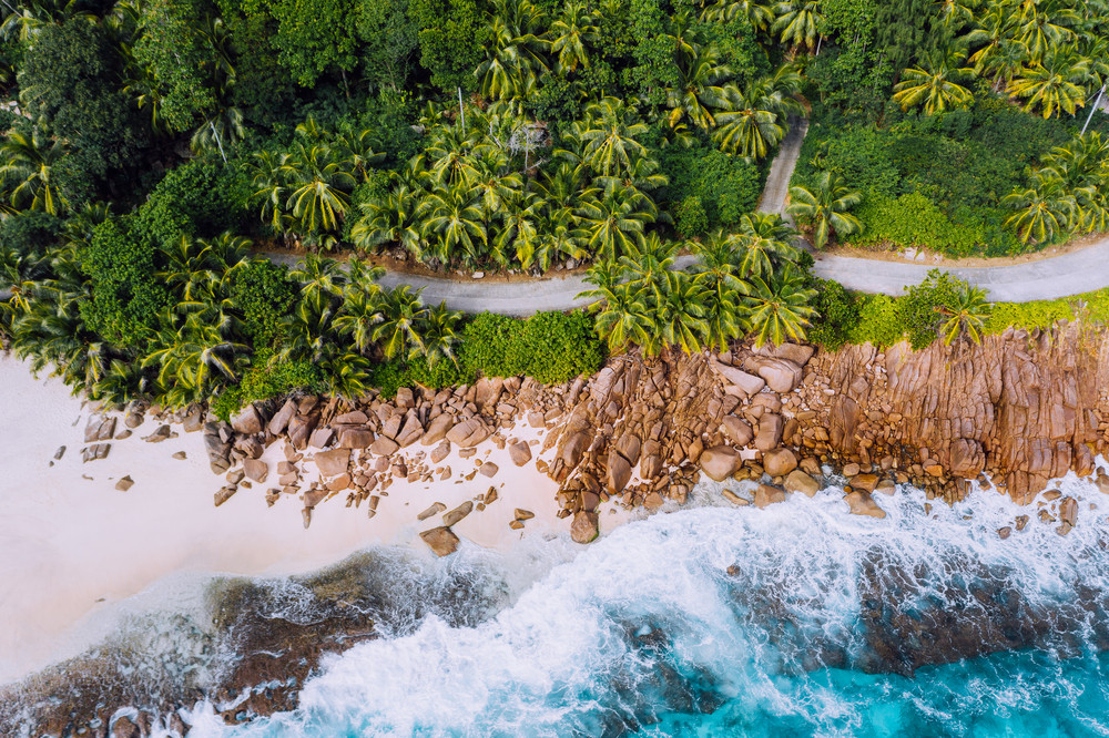 Seychelles Mahe island aerial drone landscape of coastline. Road along coastline of paradise sandy beach with palm trees and blue ocean waves rolling against granite rocks