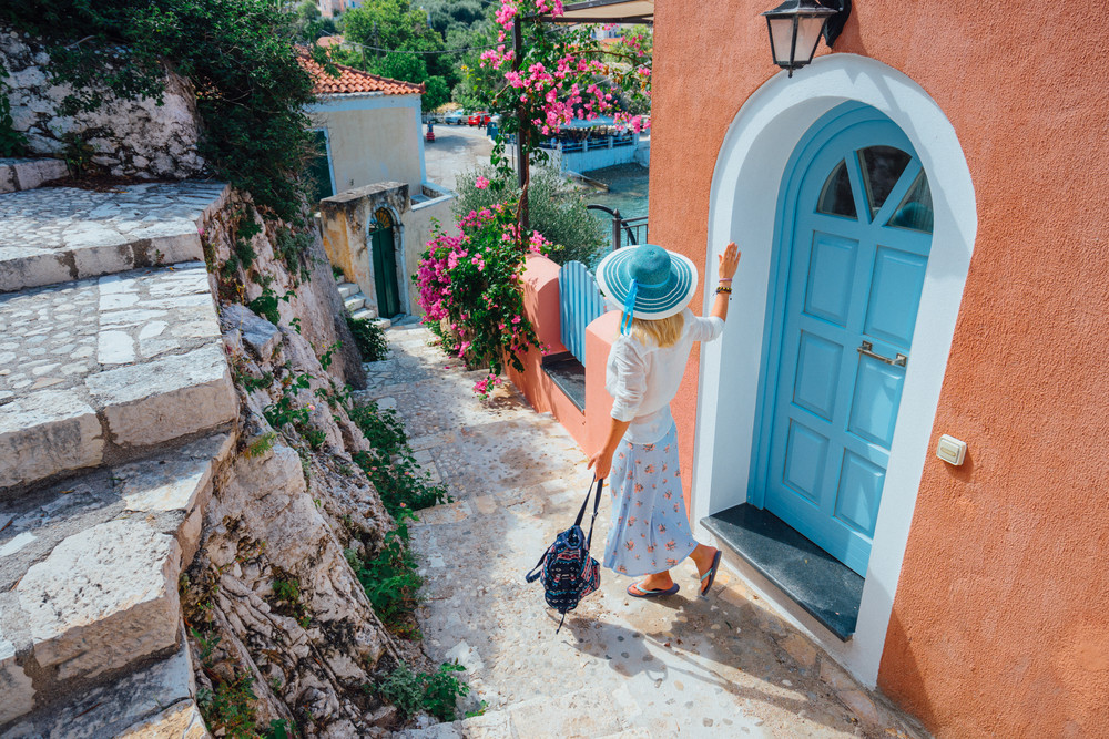 Travel tourist blonde woman with sun hat walking through narrow streets of an old greek town to the beach. Vacation summertime perfekt summer day joyful holidays fun. Greece, Europe