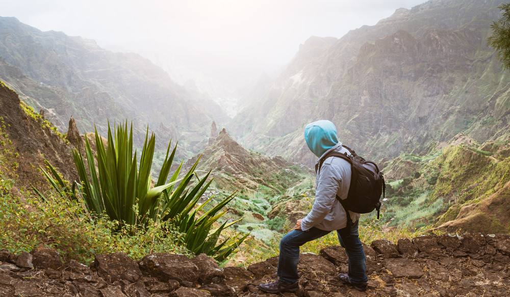 Traveler with backpack looking over the rural landscape with mountain peaks and ravine in dust air on the path from Xo-Xo Valley. Santo Antao Island, Cape Verde