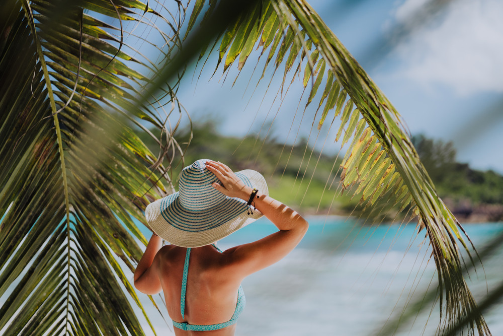 Woman on the beach in the palm trees shadow wearing blue hat. Luxury paradise recreation vacation concept